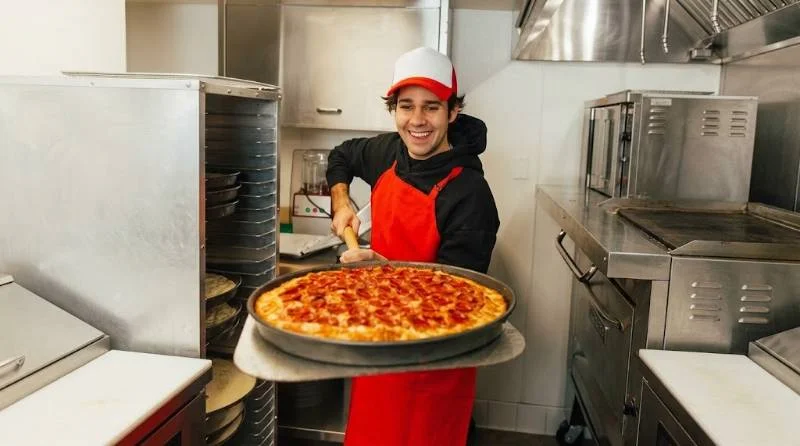 David Dobrik pulling a deep-dish pizza from an oven in a restaurant kitchen
